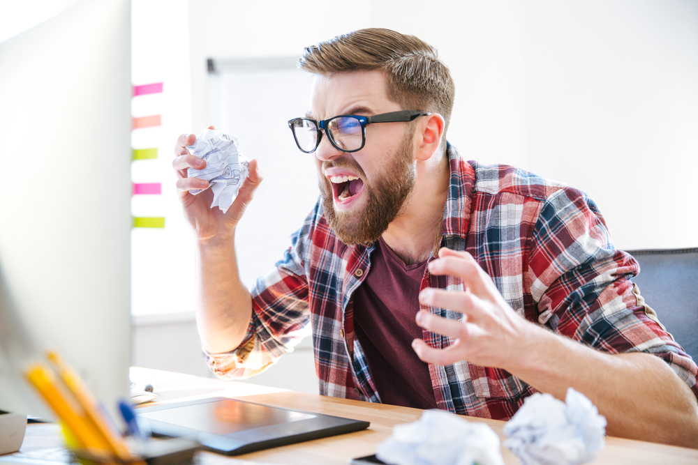 angry man sitting at a desk looking at computer with crumbled paper in his hands